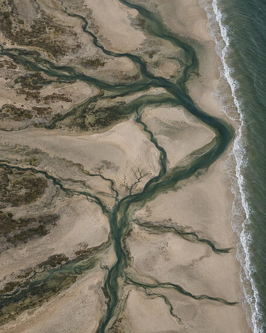 Aerial view straight down onto the Moray Firth shore at low tide, pale sand cut by dark branching tidal channels and fine ripple patterns.