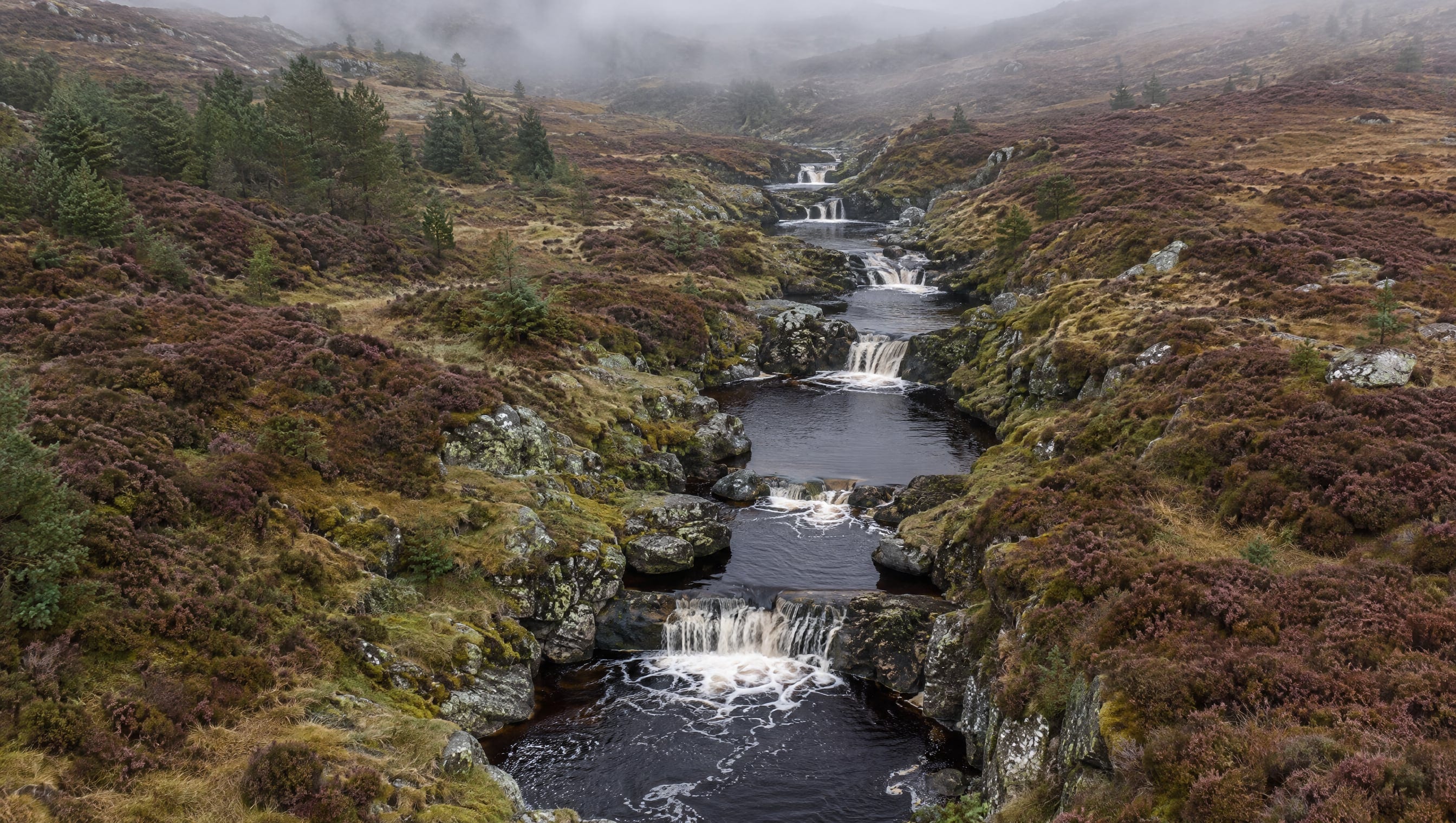 A Highland burn stepping down a heather hillside in an even sequence of small pools and falls, mist over the moor.
